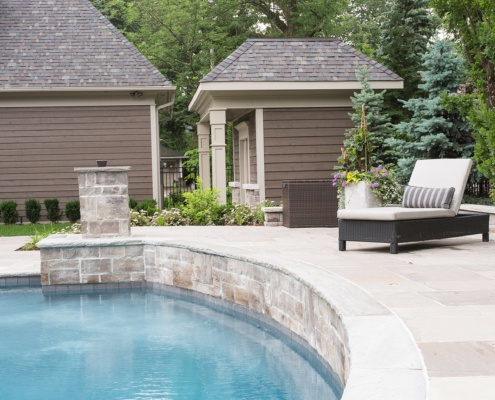 Pool house with stone skirt, shingled roof and beige trim.